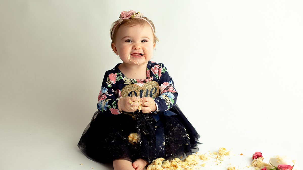 smiling toddler eats cake during one year photo session