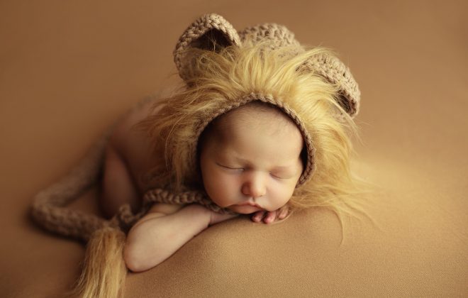 sleepy newborn posed facing forward on tan backdrop with fuzzy crocheted lion bonnet