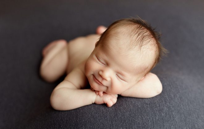 baby boy posed facing forward on gray backdrop smiles during his newborn photo session