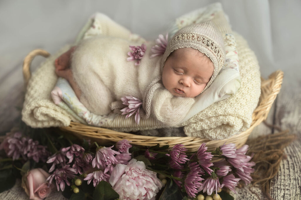 newborn baby posed on a white basket, wearing a cream knit romper and a lace cream bonnet, surrounded by purple flowers.