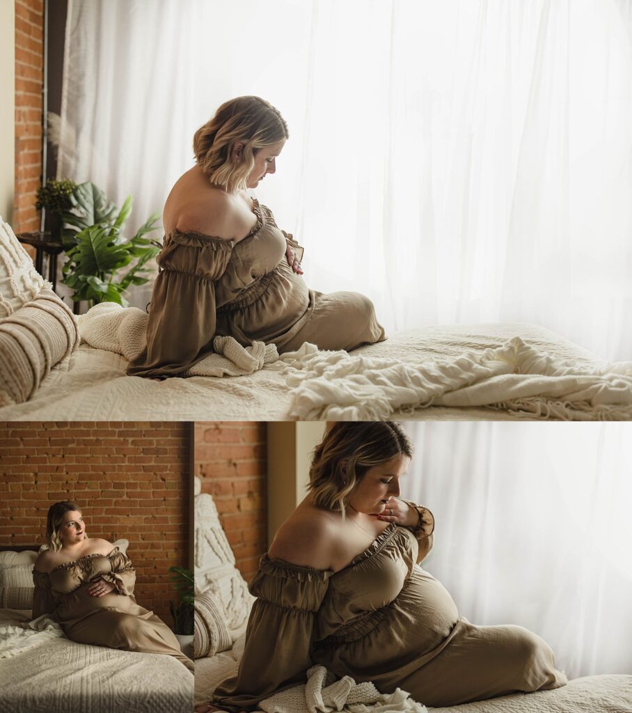 Maternity session with mother sitting on bed in a champagne colored off the shoulder silky dress, backlit by a window curtain.