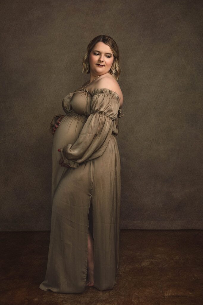 fine art style photo of an expectant mom during her maternity session, wearing a champagne off the shoulder silky dress and gazing down over her shoulder.