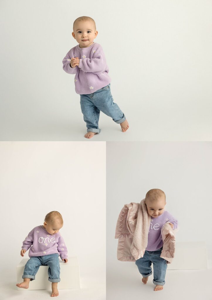 A one year old in a purple sweater and jeans against a white backdrop.  She is smiling while trying to run away from the camera.