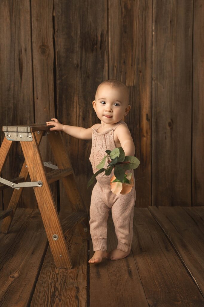 A one year old in pink knit overalls leaning on a small vintage ladder and holding a flower toward the camera.