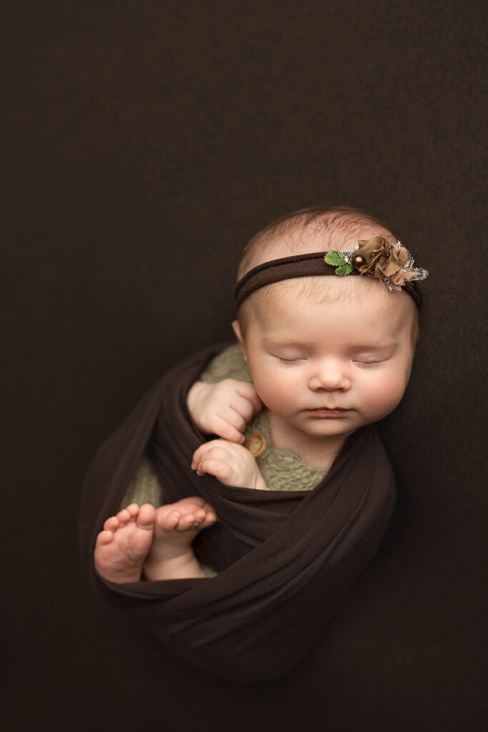 newborn baby wrapped in a brown wrap and posed on a matching brown backdrop.