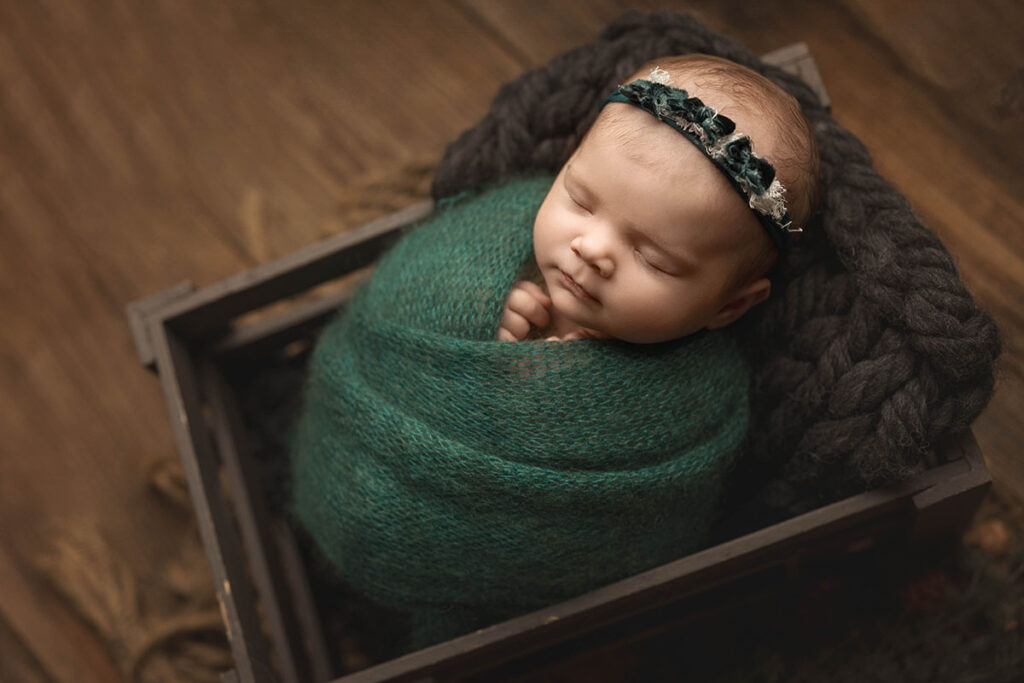 sleeping newborn baby wrapped in a jewel toned green wrap, laying in a gray crate.