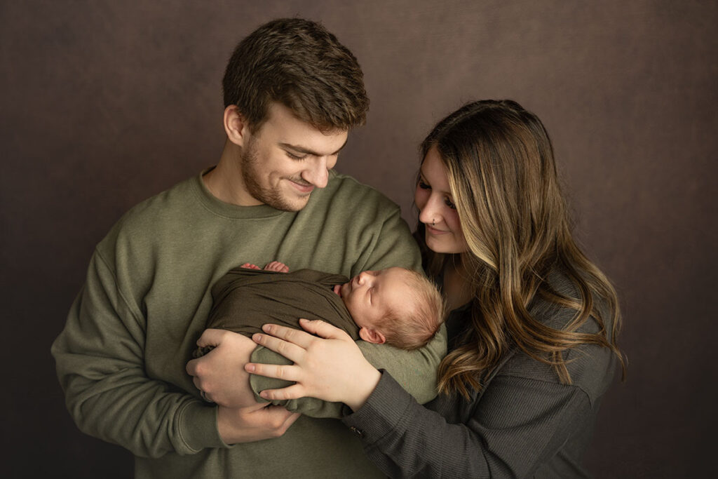 Dad holding newborn baby and smiling down at him, while mom leans over dad's shoulder to gaze at her new baby.