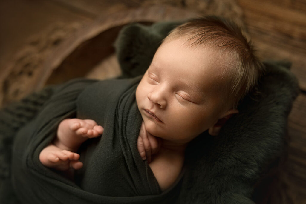 Newborn baby wrapped with a forest green wrap and laying in a rustic wooden bowl for his newborn photos.