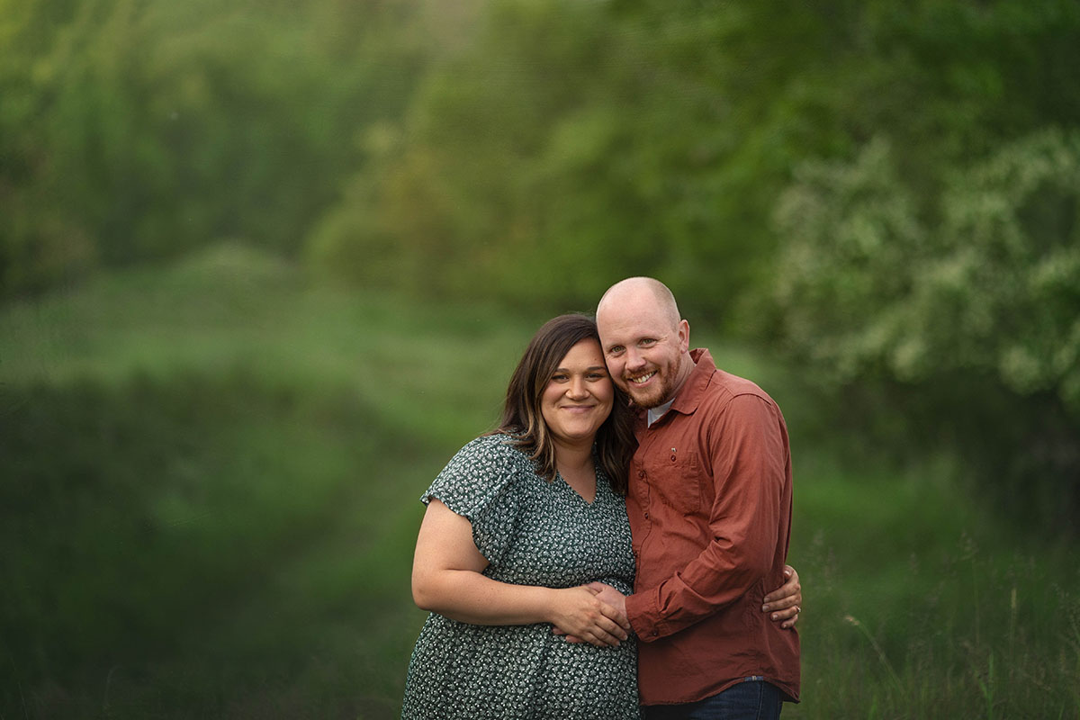 expectant mom and her partner smile at the camera with their arms around each other.  Softly focused green leafy trees are in the background.