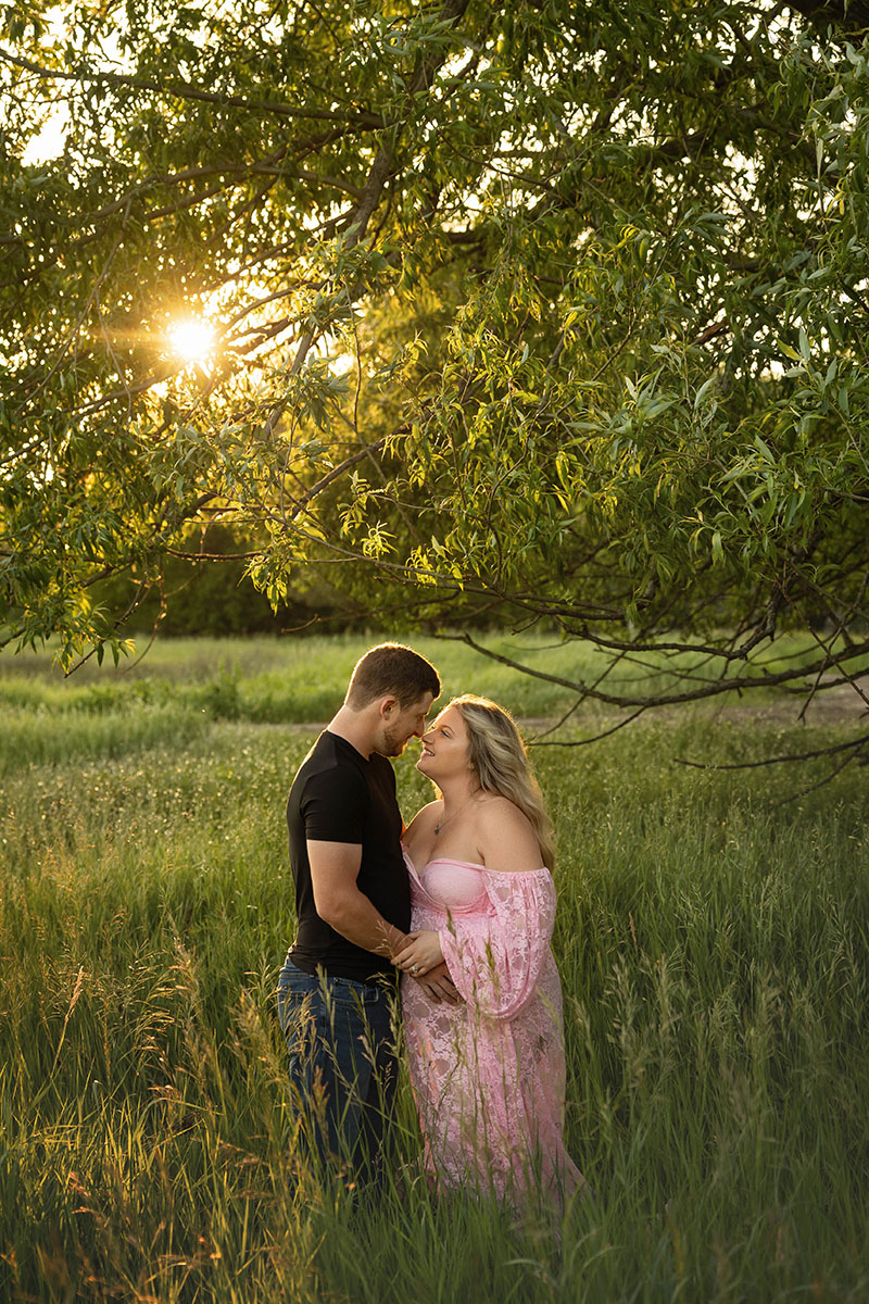 expectant mom wearing pink maternity dress gazes at her partner. They stand under a tree with gorgeous golden backlight flooding behind them.