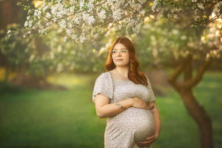whimsical maternity portrait with expecting mom hugs her belly and gazes into the distance. Golden light and apple tree blossoms are soft in the background.